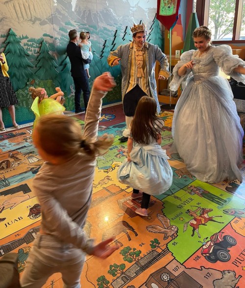 Children dance joyfully around a prince and princess in a colorful, playful setting with forest-themed decor.