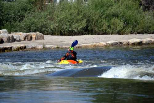 A kayaker in an orange kayak paddles through a river with rocky banks and lush greenery in the background.