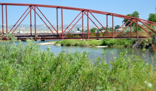 A red metal bridge spans a calm river, surrounded by lush greenery and distant hills under a clear blue sky.
