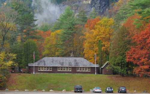 A rustic building surrounded by vibrant autumn foliage and parked cars, set against a mountainous backdrop.