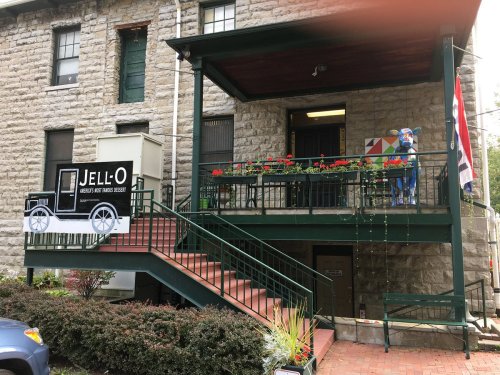 A historic building with a green porch, stairs, and a Jell-O museum sign, surrounded by flowers and colorful decorations.