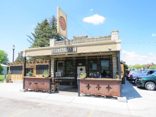 A cozy restaurant with a stone facade, outdoor seating, and a sign, set against a clear blue sky.