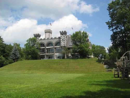 A large stone castle with turrets sits atop a grassy hill, surrounded by trees under a partly cloudy sky.