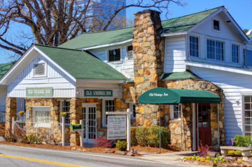 A charming inn with a stone facade and green awnings, surrounded by trees and flowers on a sunny day.