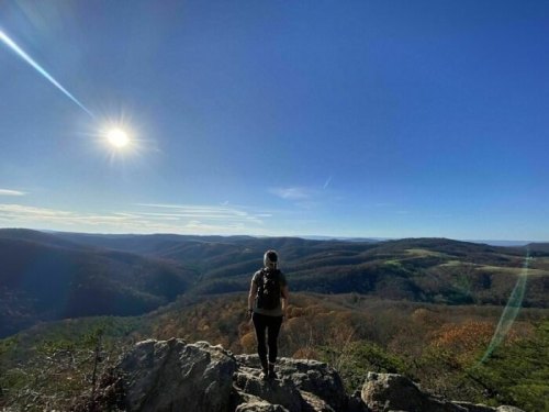 A person stands on a rocky outcrop, gazing at a vast landscape under a clear blue sky with the sun shining brightly.