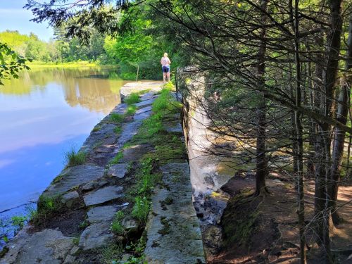 A person stands on a stone ledge by a calm river, surrounded by trees and greenery, with a small waterfall nearby.