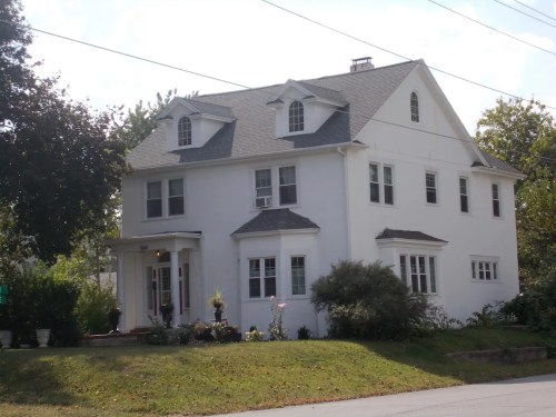 A large white two-story house with a sloped roof, surrounded by greenery and flower beds.