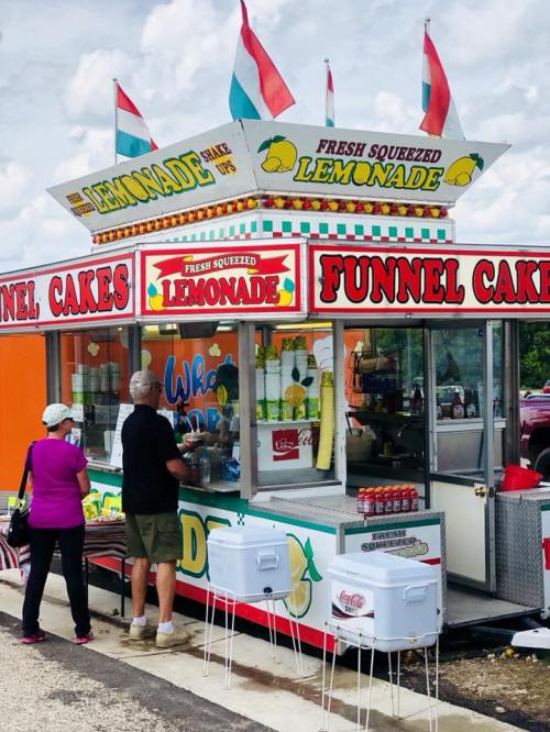 A colorful food stand selling funnel cakes and fresh squeezed lemonade, with customers ordering at the counter.