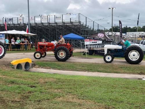 Two vintage tractors drive along a path at an outdoor event, with spectators and tents in the background.