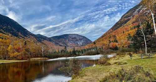 Scenic view of a tranquil lake surrounded by autumn-colored mountains and trees under a blue sky with clouds.
