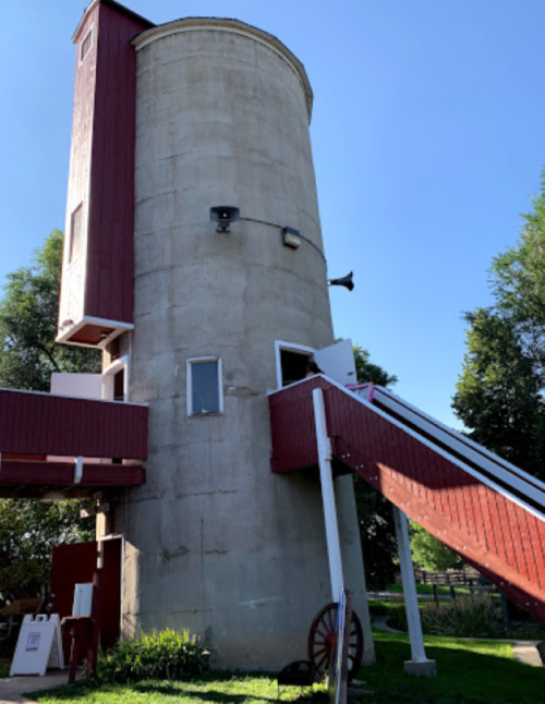 A tall, cylindrical concrete silo with a red wooden addition and a staircase leading to a door. Green trees surround it.