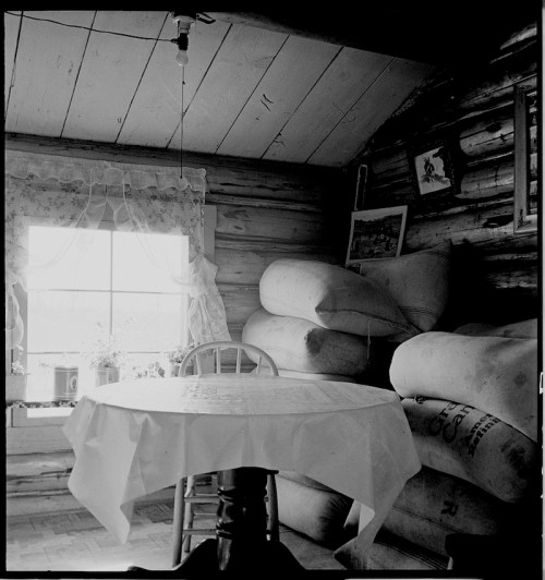 A cozy, rustic room with a table, stacked bags, and a window adorned with lace curtains.