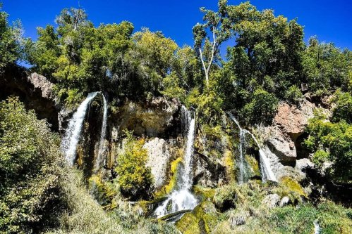A scenic view of cascading waterfalls surrounded by lush green trees and a clear blue sky.