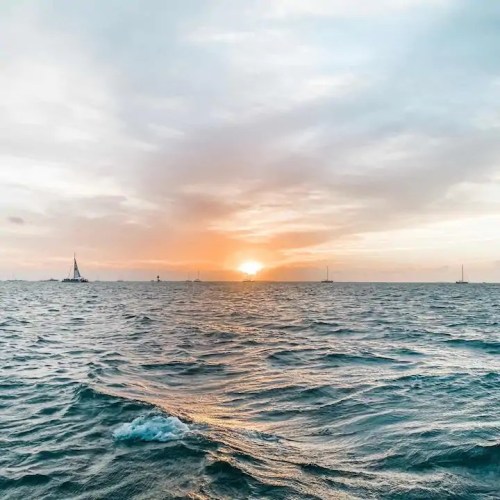A serene seascape at sunset, with gentle waves and distant sailboats against a colorful sky.