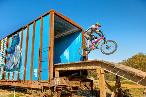 A cyclist jumps off a ramp from a colorful train car, performing a trick against a clear blue sky.