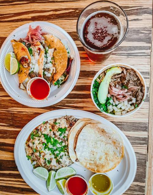 A table with two plates of tacos, a bowl of meat, and a glass of beer, garnished with lime and sauces.