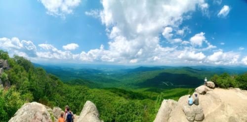 Panoramic view of lush green mountains under a bright blue sky with fluffy clouds, featuring hikers on rocky outcrops.