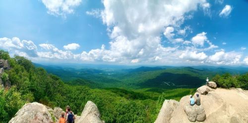 A panoramic view of lush green mountains under a partly cloudy sky, with hikers on rocky outcrops.