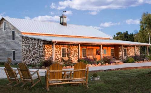 A rustic stone house with a metal roof, surrounded by greenery and featuring wooden chairs in front.