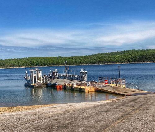 A ferry docked at a calm lake, surrounded by green trees under a clear blue sky.