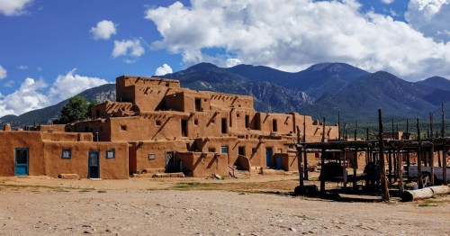 A traditional adobe building with blue doors, set against a backdrop of mountains and a partly cloudy sky.