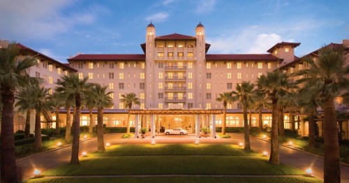 A grand hotel with palm trees, illuminated at dusk, featuring a welcoming entrance and elegant architecture.