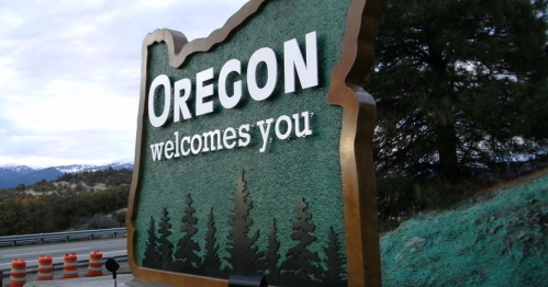 Sign welcoming visitors to Oregon, featuring the state outline and trees, with mountains in the background.