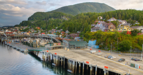 A scenic waterfront view of a small town with colorful buildings, boats docked, and lush green hills in the background.