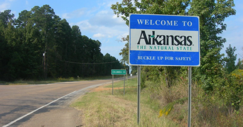 Welcome sign for Arkansas, "The Natural State," with a reminder to buckle up for safety, alongside a rural road.