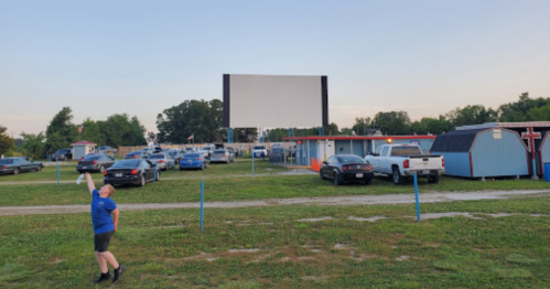 A person holds a drink in front of a drive-in theater with parked cars and a large blank screen in the background.