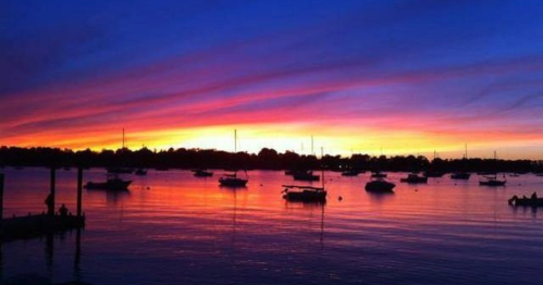 A vibrant sunset over a calm waterway, with silhouettes of boats and colorful clouds reflecting on the surface.