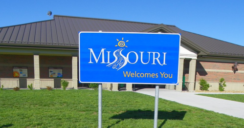 A blue welcome sign for Missouri in front of a building, with green grass and a clear blue sky in the background.