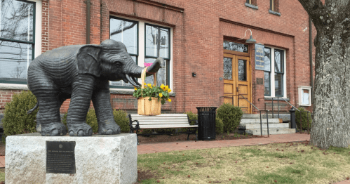 A bronze elephant statue holding a basket of flowers in front of a brick building with large windows.