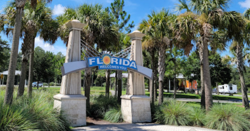 A welcoming sign for Florida surrounded by palm trees and greenery under a clear blue sky.