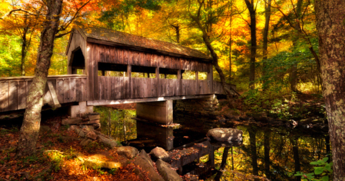 A wooden covered bridge over a serene stream, surrounded by vibrant autumn foliage.