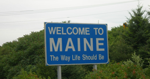 A blue welcome sign for Maine, stating "Welcome to Maine - The Way Life Should Be," surrounded by greenery.