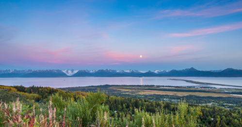 A scenic view of mountains and a calm lake at sunset, with soft pink and blue skies above.
