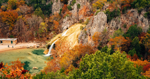 A scenic view of a waterfall cascading into a clear pool, surrounded by vibrant autumn foliage and rocky cliffs.