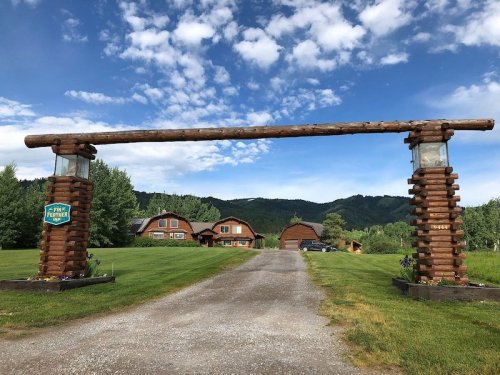 A rustic wooden entrance gate leads to a scenic property with a large house and green landscape under a blue sky.