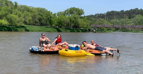 Four people relax on inflatable rafts in a river, surrounded by greenery and a bridge in the background.