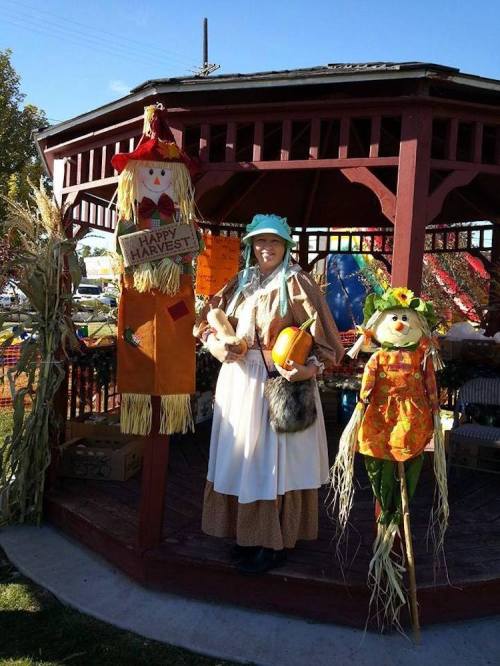 A woman in a historical costume stands beside two colorful scarecrows in a gazebo decorated for harvest.