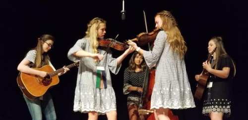 Five young musicians perform on stage, playing guitar, violin, and cello, dressed in casual outfits.