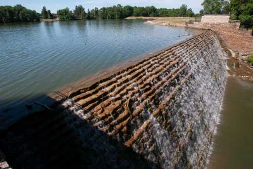 A stone dam with water cascading over its edge into a calm lake, surrounded by greenery and trees.