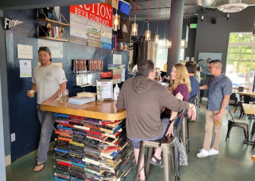 A cozy bar with a book-themed counter, patrons chatting, and a bartender serving drinks in a casual setting.
