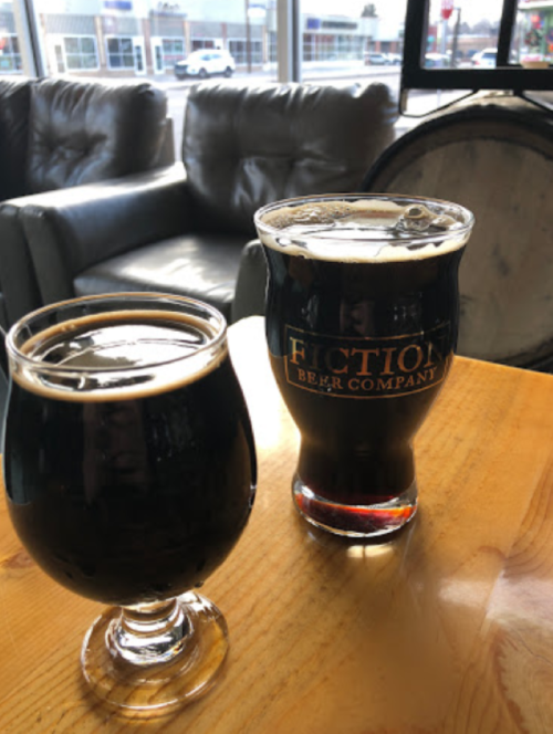 Two dark beers in glasses on a wooden table, with a cozy seating area in the background.