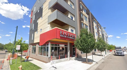 Exterior view of Fiction Beer Company, a modern building with red awning, surrounded by trees and clear blue sky.