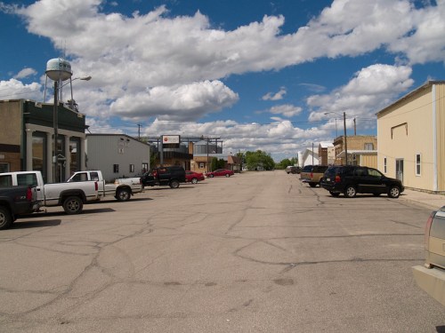 A wide street in a small town with parked cars, buildings on either side, and a blue sky with fluffy clouds.