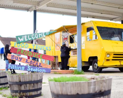 A colorful sign welcomes visitors to a food truck area with a yellow truck and planters in the foreground.
