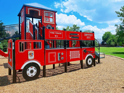 A colorful playground structure designed like a fire truck, featuring slides and climbing areas, set in a grassy park.