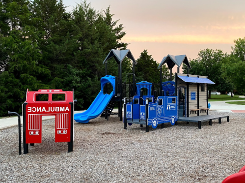 A colorful playground featuring a red ambulance structure, blue slides, and a playhouse, surrounded by trees.